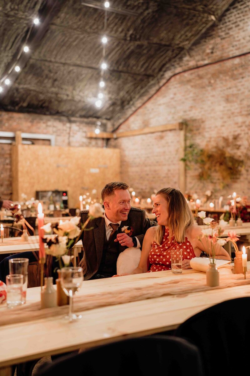 A candid photograph of two wedding guests. They are a couple and sit alone in a large Aberdeenshire barn. They sit in a room that's been set up for dinner and the moody glow of candles can be seen all around.