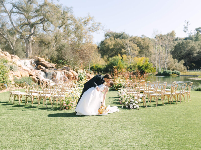 Bride and groom share a romantic kiss in a sunlit garden, surrounded by decorative chairs and vibrant flowers, with a waterfall in the background.