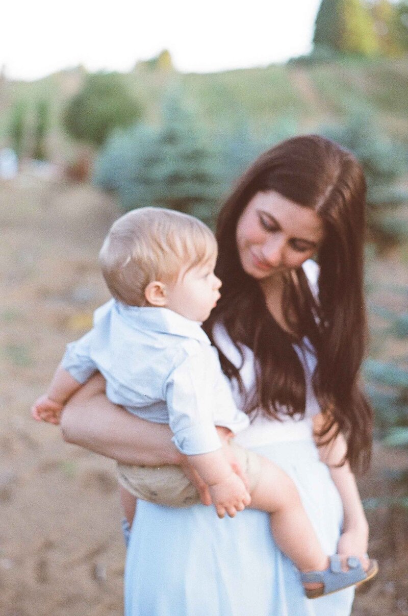 Husband wearing white dress shirt and khaki pants,  wife wearing blue flowy long dress and blonde curled hair, and two year old son wearing white dress shirt and khaki shorts walking away from view walking down a dirt pathway lined with wildflowers on mountainside ledge by Portland Newborn Photographer Emilie Phillipson Photography.