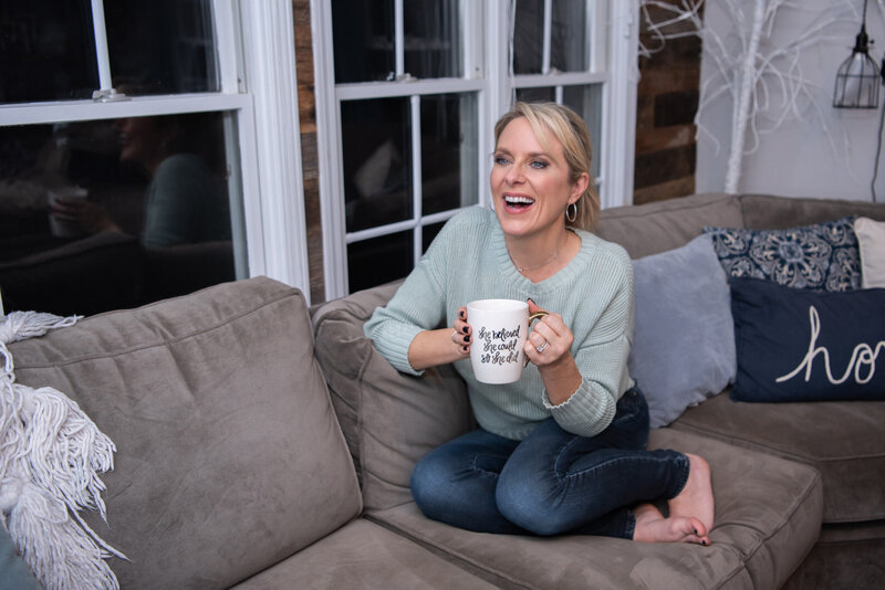 A woman is laughing and sitting on a couch while holding a mug.