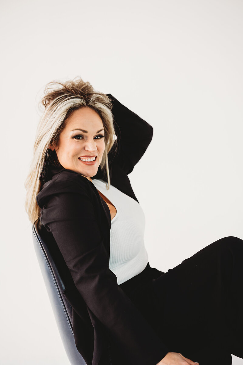 Woman in business suit sitting in a chair against a white wall and smiling