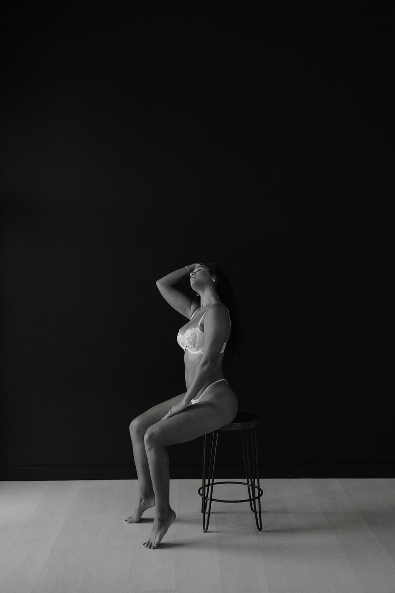 Black and white photo of woman in white lingerie sitting on a stool in front of black wall
