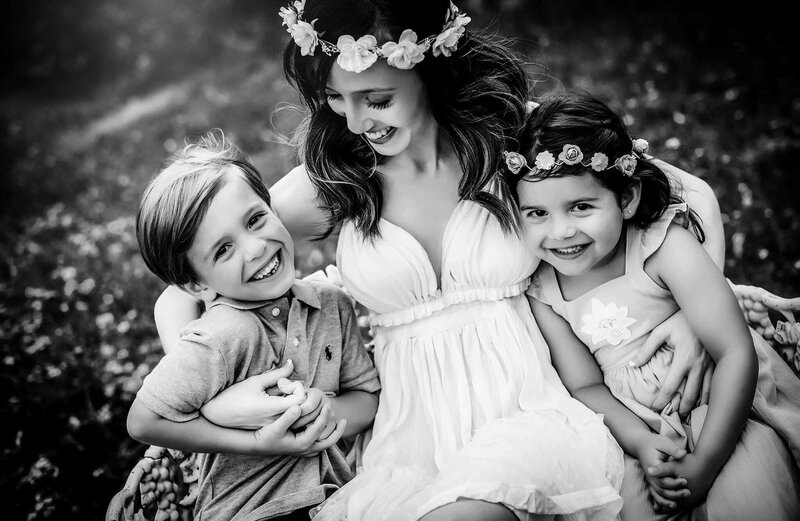 A black and white photo captures a woman with two young children, all smiling under the sun. The woman and girl wear floral headpieces, while the boy sports a polo shirt. The serene setting suggests a meadow or park, showcasing the tender artistry characteristic of Bia Schaefer Photography.