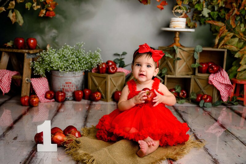 little one year old girl sits in her custom cake smash set up "apple of my eye" during her milestone session in a waxhaw studio.  she is wearing a red bow and red dress