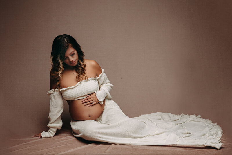 Maternity portrait of dark curly haired woman in white long sleeve crop top and white skirt sitting on floor holding belly and looking at baby bump