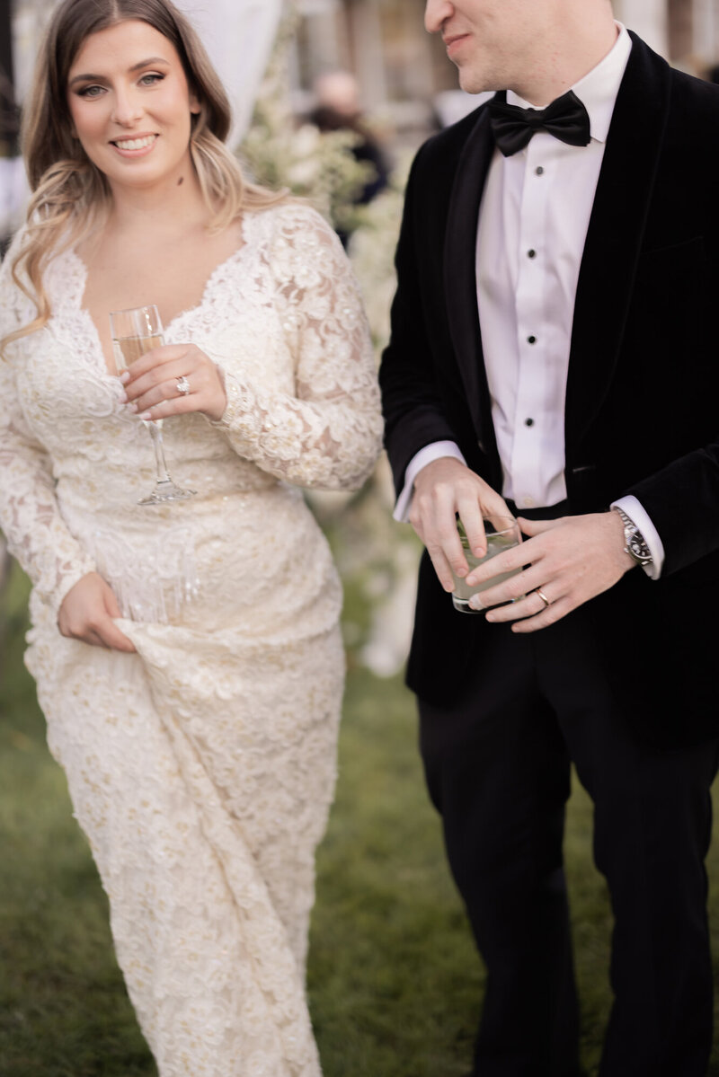 Bride and groom hold drinks and smile during their outdoor wedding reception in the Hamptons, with the bride in a lace gown and groom in a black velvet tuxedo.
