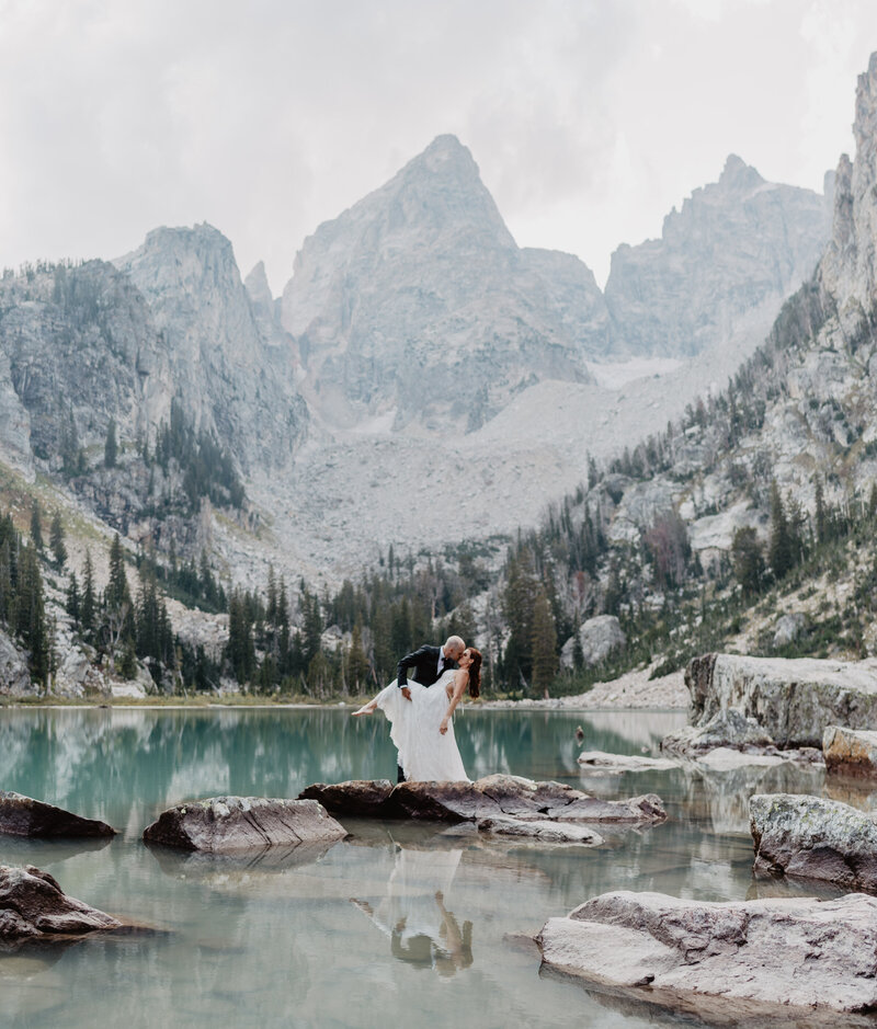 Jackson hole wedding  captures Delta Lake wedding pictures in Wyoming with bride and groom kissing passionately while standing on a rock together in front of the Tetons