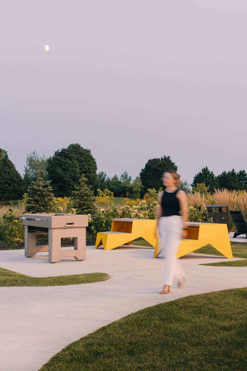 A woman walking along a concrete pathway through an outdoor game area, featuring a foosball table, two bright yellow chairs, and lush greenery