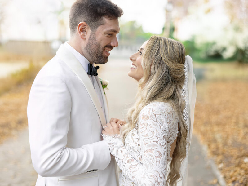 A bride and groom stand closely, smiling at each other. The bride wears a white lace dress with long sleeves and a veil, while the groom is in a white suit with a black bow tie. They are outside on a path lined with autumn leaves.