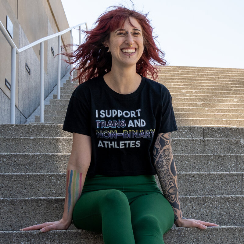 a woman sits outdoors on steps in a shirt that supports trans and non-binary athletes to show your inclusive strength journey