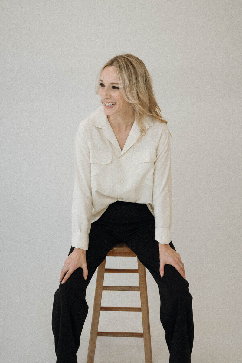 Joy Steele, Phoenix boudoir photographer, smiling while seated on a wooden stool in a minimalist studio wearing a white blouse and black pants.