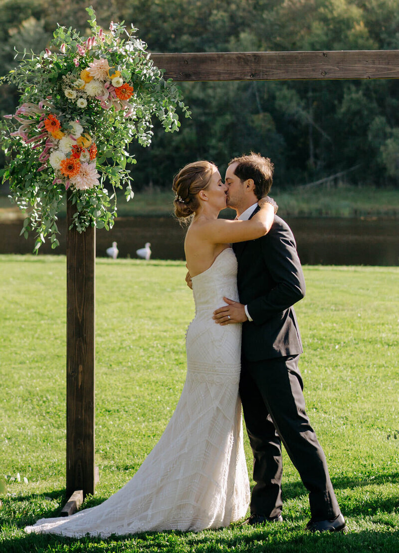 The groom and bride are kissing near the altar during the wedding ceremony by the river.
