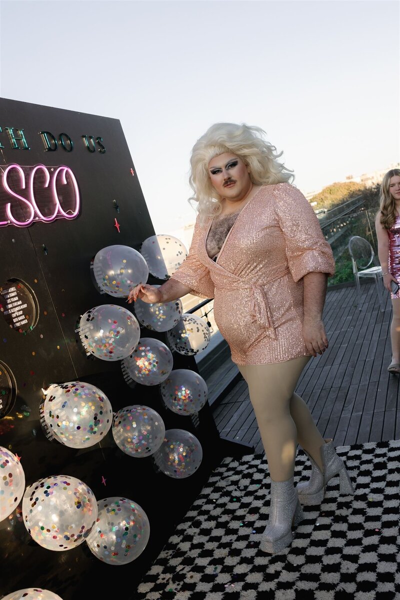 Caucasian mustached drag queen with blonde long haired wig in blush sequin romper smizing at the camera while popping a confetti filled balloon escort wall on a black and white checkered shag rug on the terrace at the Line Hotel DC 