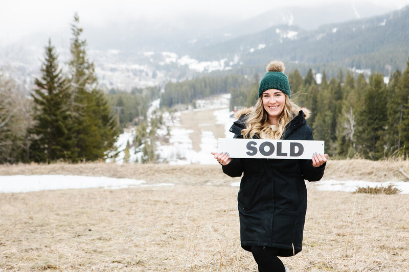 Realtor Tamara Rzan proudly holding a 'Sold' sign against the majestic mountain backdrop of the Crownest Pass