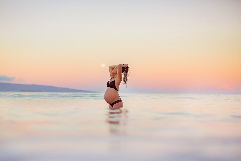 Woman in black bikini holding hair and closing eyes during pregnancy portraits in the ocean