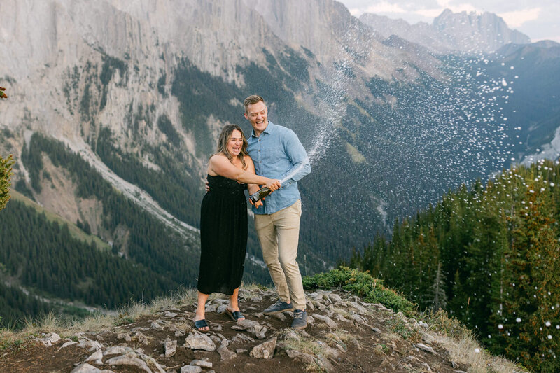 Capture the joyous moment of a couple popping champagne and celebrating their new engagement on top of a mountain after a hiking adventure. A thrilling and romantic scene set against the breathtaking backdrop of the Rockies.