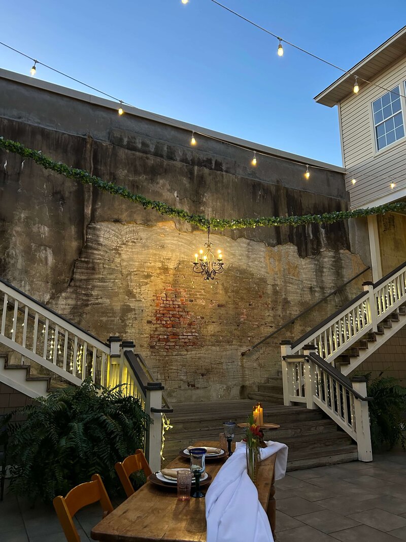An outdoor scene with a brick and plaster wall, a chandelier, a wooden table set with flowers, candles, and tableware, green plants, and a white staircase decorated with lights.