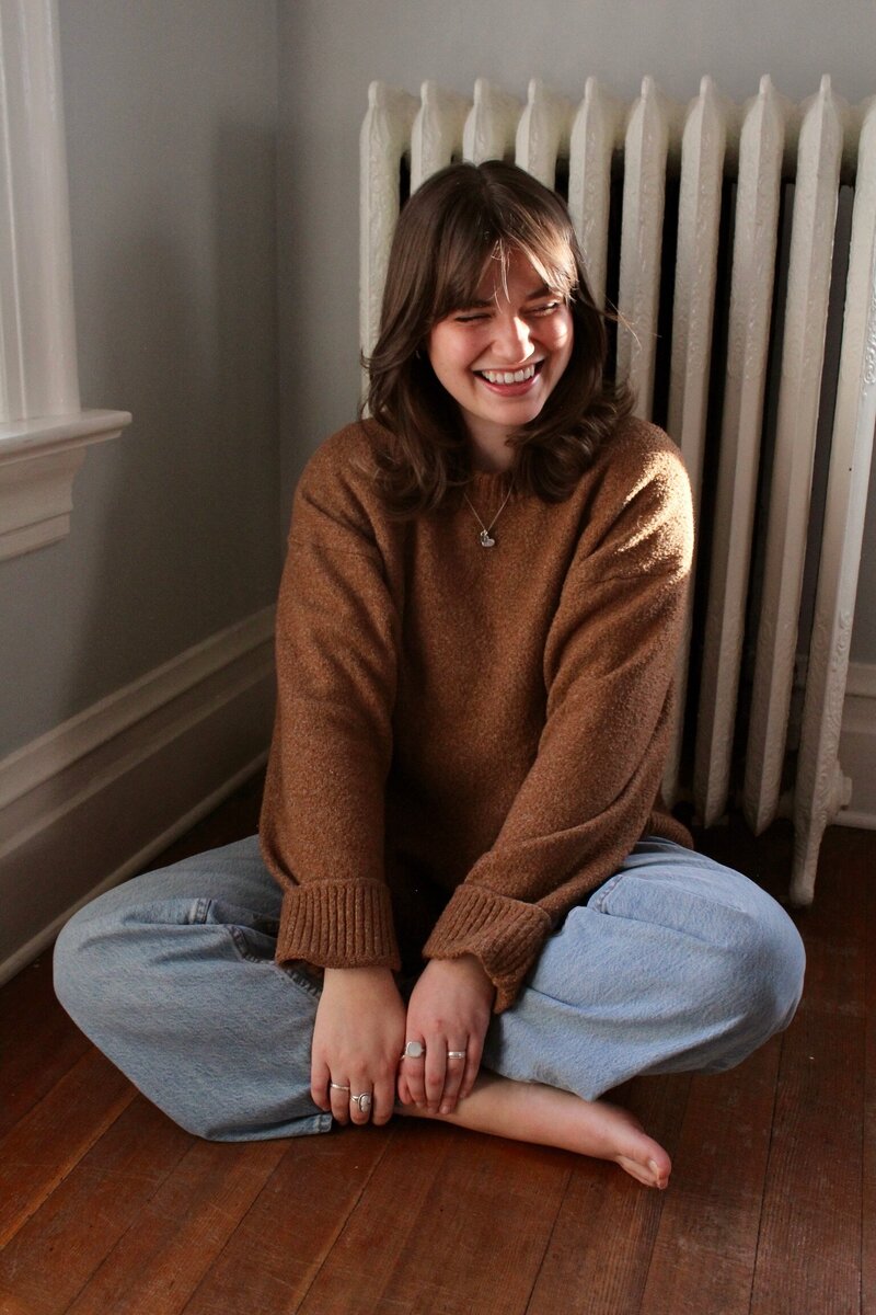 Woman wearing a brown sweater and jeans sitting on the floor in front of a radiator with her legs crossed and laughing