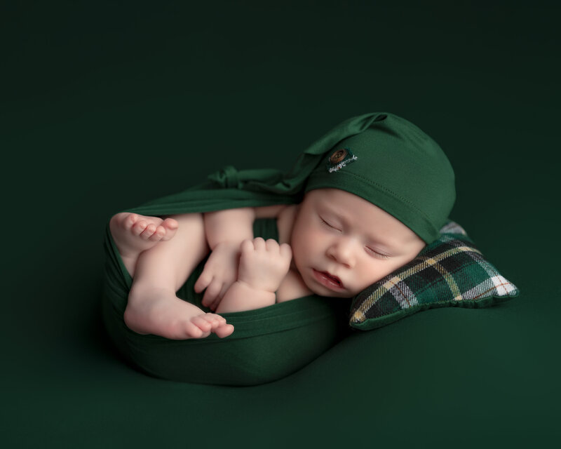 Cozy newborn portrait of a baby boy in a wrapped Huck Finn pose with plaid pillow, styled in rich greens – Paris, Ontario
