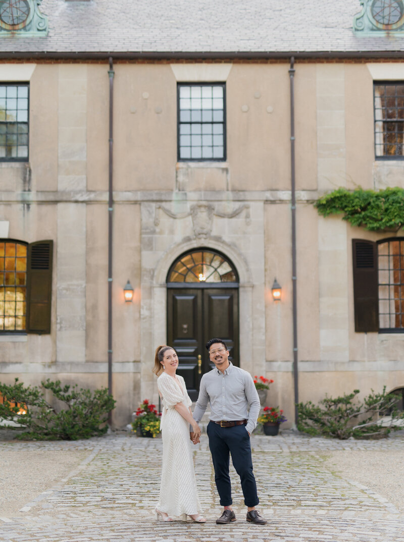 photographer and creative director kaylyn leighton holds husband hand in front of beautiful estate for an engagement session. Photographer, Joe sinthavong laughing at camera on a late summer day.