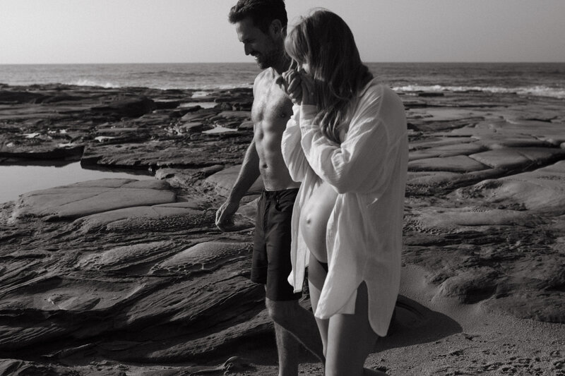Couple walking hand inhand , the pregnant woman holds her hand up to her lips and kisses hs hand. Black and white image they are on a beach , she is wearing a white button up shirt with exposed pregnant belly