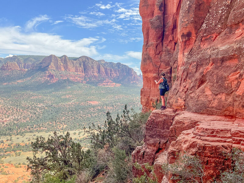 A woman is standing on a cliff in Sedona, Arizona taking a picture of the landscape during a hike