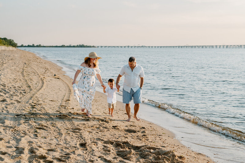 Family of three walking on the beach in white next to the ocean in Kent Island Maryland.