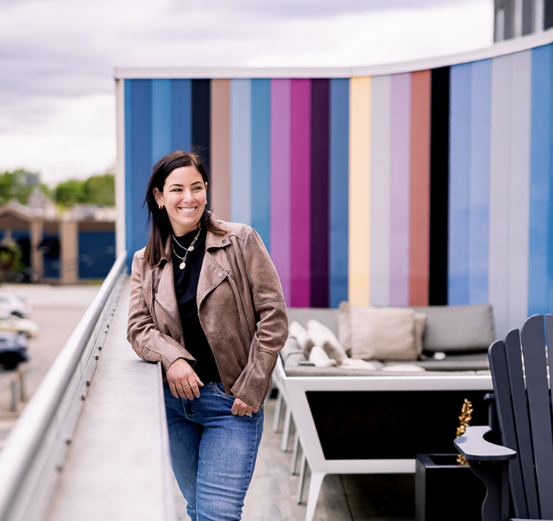 Becky Reel smiling in a brown leather jacket leaning against a wall and looking to her left with a colorful wall background