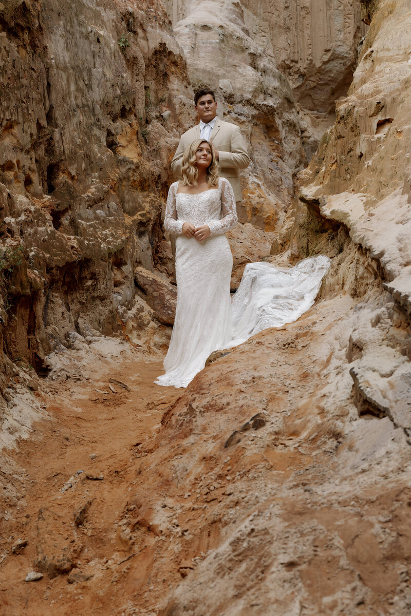 An eloped couple stands among the red rocks with the bride in the front, while taking in their elopement ceremony location.