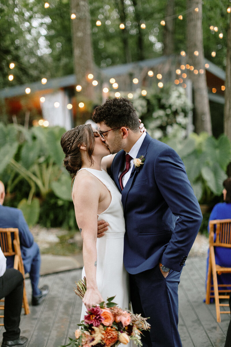 A couple embraces during their wedding ceremony, surrounded by greenery and warm string lights, with a floral bouquet in hand.
