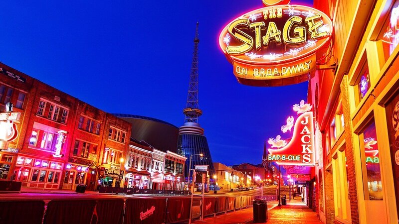 A vibrant nighttime view of Nashville's iconic Broadway street, illuminated by colorful neon signs for bars, restaurants, and live music venues, with the blue sky and modern architecture in the background. This energetic scene reflects the lively cultural adventures supported by vacation fundraising for charities.