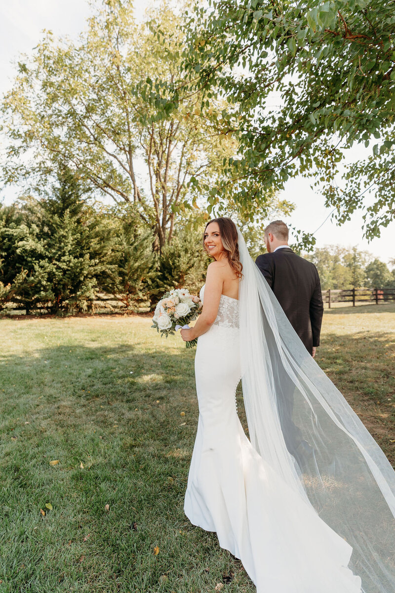 A bride in a white gown and long veil holds a bouquet while walking with a groom in a dark suit at an outdoor wedding venue in Virginia. They stroll through a grassy area, trees, and wooden fence behind them; the scene captures the essence of barn wedding pictures as she looks back smiling.