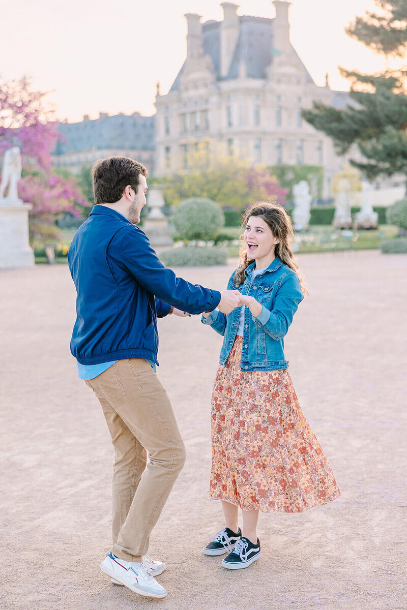 “Man and woman holding hands during a Paris proposal at the Louvre and Tuileries Garden — the man smiling as the woman looks on in excitement.