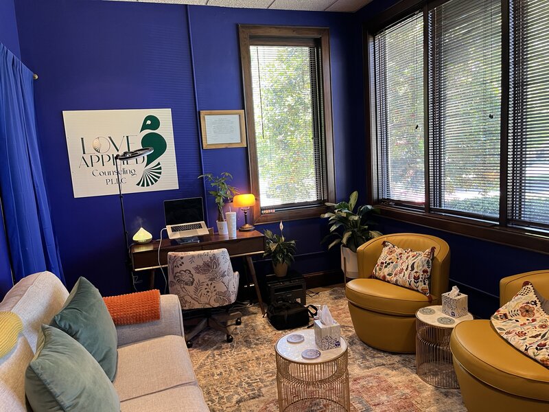 The interior of the Love Applied Counseling office is  dark green, with an art deco aesthetic and gold accents. A pink-patterened rug is centered in the room, with  two comfortable chairs and a modern office desk along the office walls. The space is calming and comforting, with plants, colorful paintings, and a tea cart.