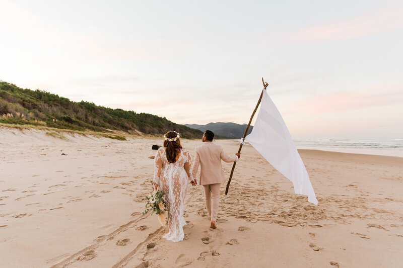 Wedding couple walking down beach at sunset with flag