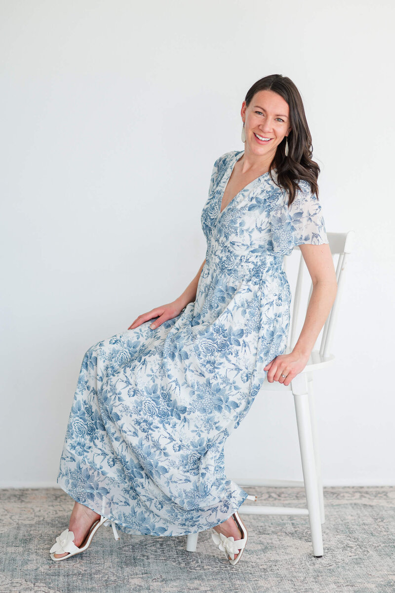 Indoor studio portrait of woman in white and blue dress sitting on a white chair against a white background.