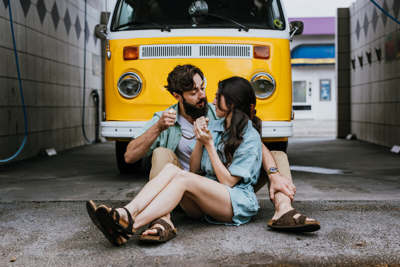 Couple sits on ground in front of yellow VW bus at car wash