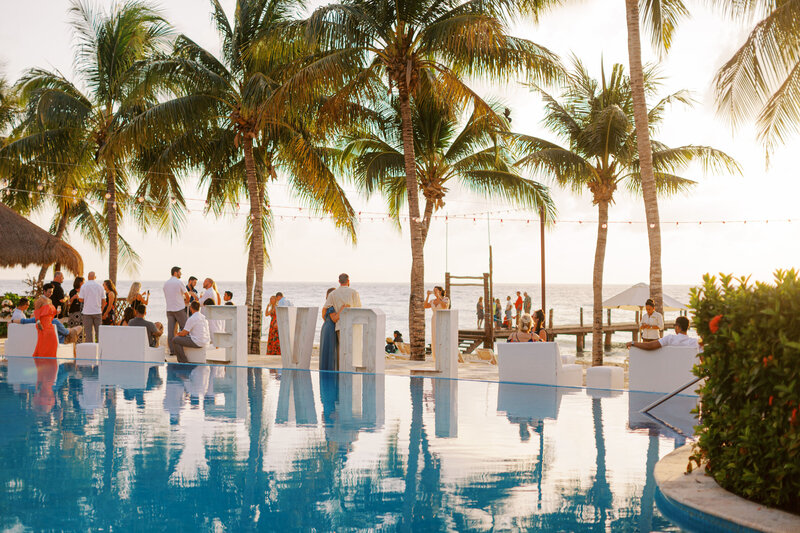 Beachfront resort pool at sunset with palm trees and guests enjoying the view.
