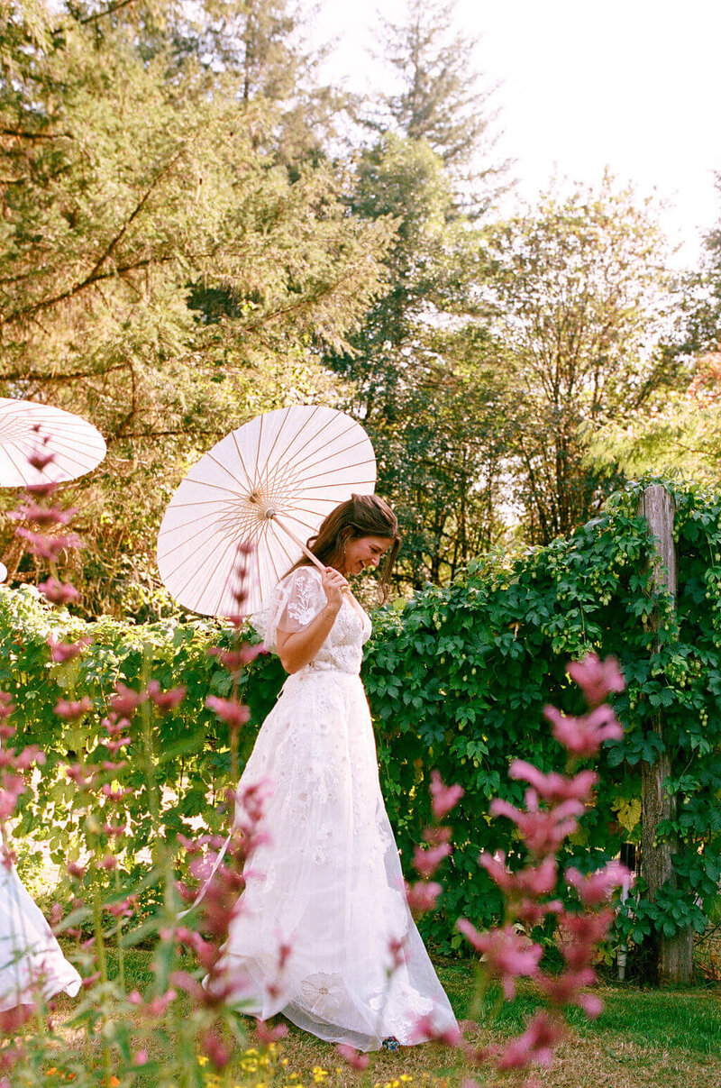 A bride holds a parasol and walks in a flower garden, representing the event coordinator services of Birdie June Events and Rentals. 