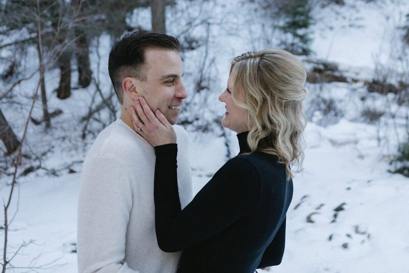 A woman holds the face of her fiance as they look at each other and smile with a snowy backdrop.