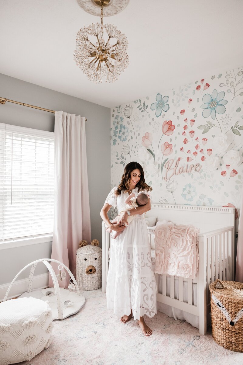 A woman standing in a nursery, tenderly holding a newborn, beside a crib with pastel bedding and a floral wall design, captured during an at-home newborn photography session.