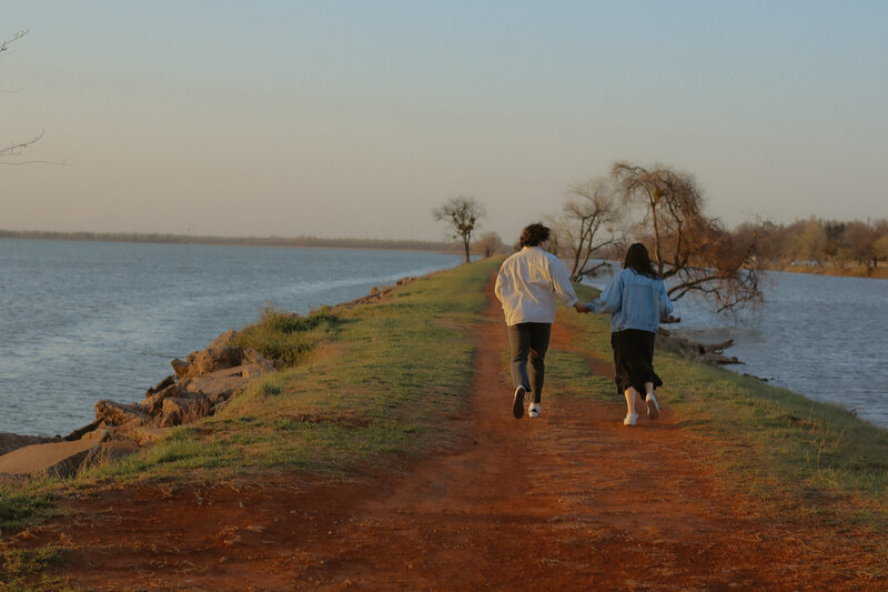 Man and Woman holding hands while running away from the camera. They are on a strip of land with water on wither side of them.