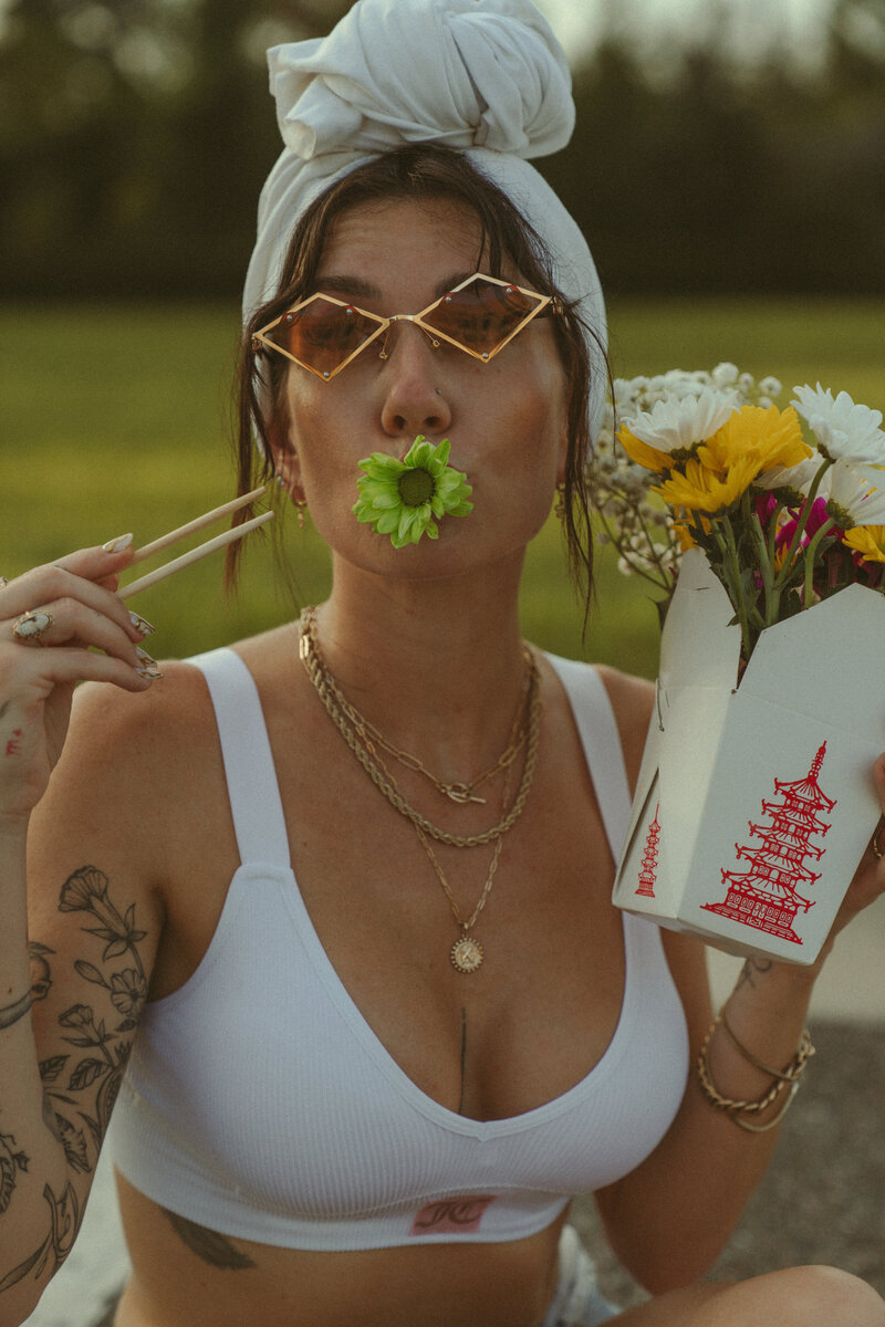 Stylish woman wearing gold geometric sunglasses, white head wrap, and layered gold jewelry, holding a flower in her mouth and Chinese takeout box filled with colorful flowers in a playful editorial-style portrait outdoors.