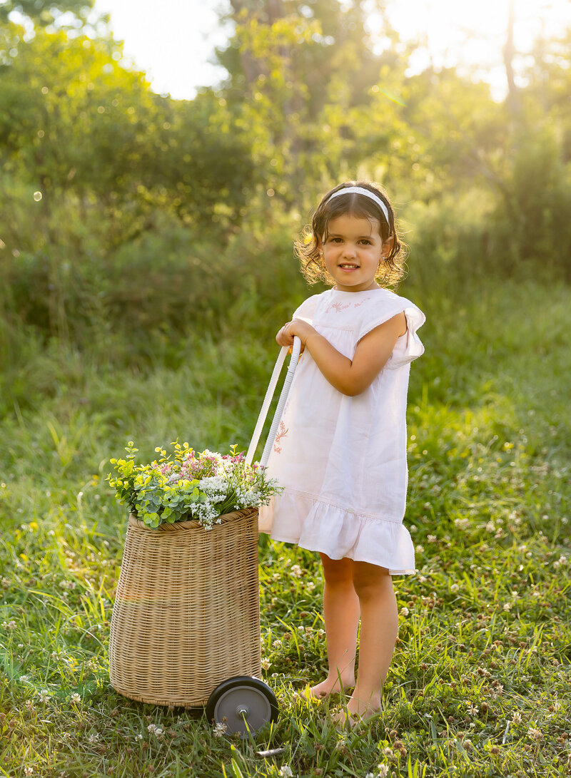 Little girl is standing barefoot in a field with a flower basket. The is surrounded by soft golden light. She is wearing a white dress and smiling at the camera. Captured by best Brooklyn, NY family photographer Chaya Bornstein.