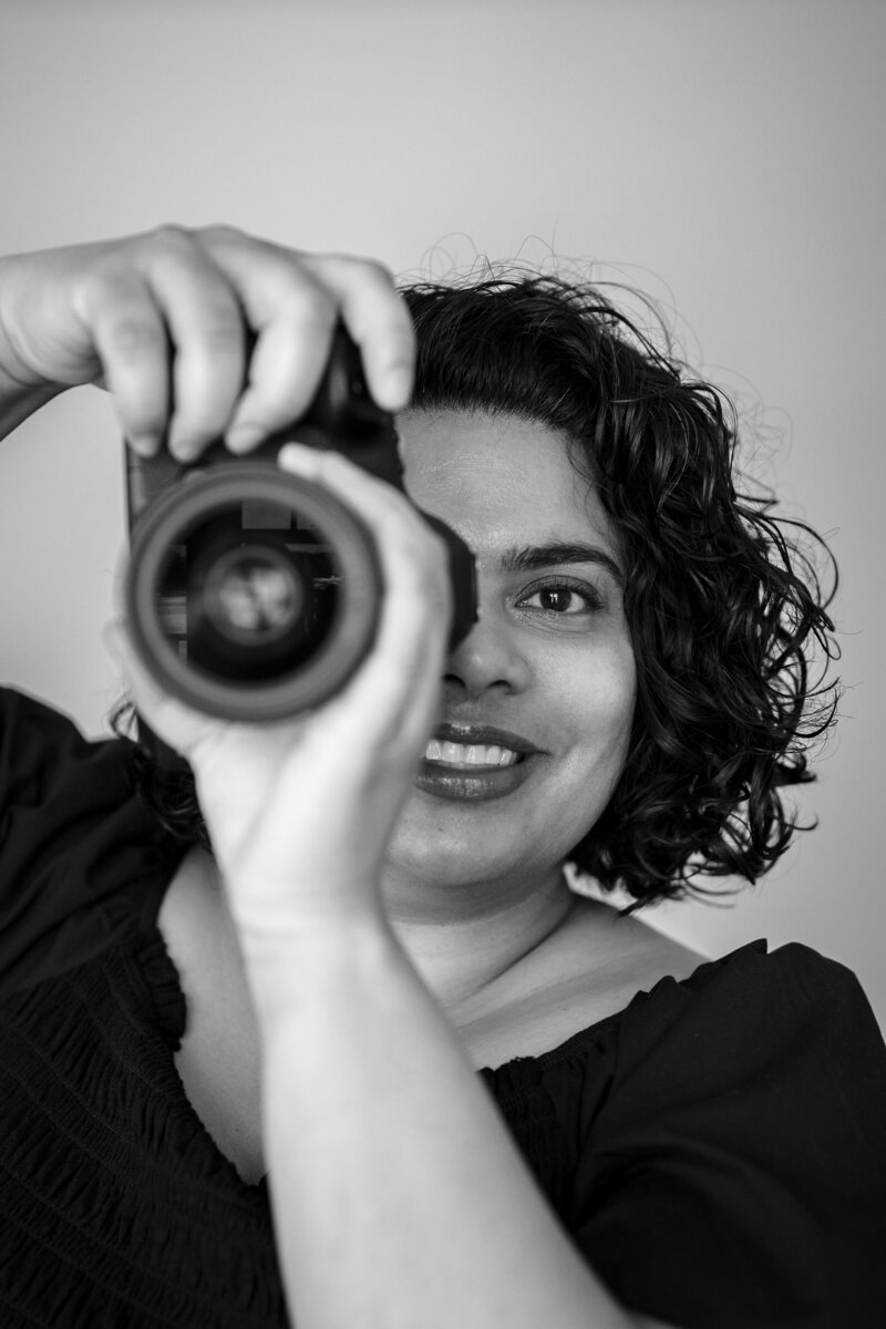 A black and white photo of a woman holding up a camera, smiling as she looks through the viewfinder.