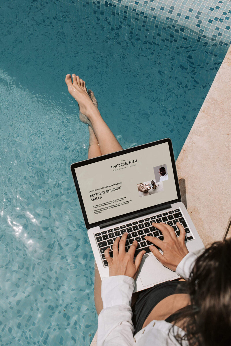 A women by the pool with The Modern Life Coach School on her  laptop