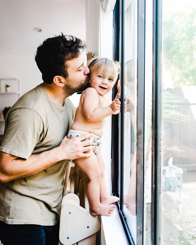 Dad kissing baby girl who is standing in a window at home