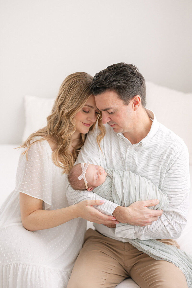 Mom and dad snuggling close as dad holds their newborn baby girl on a white bed in a bright, white studio