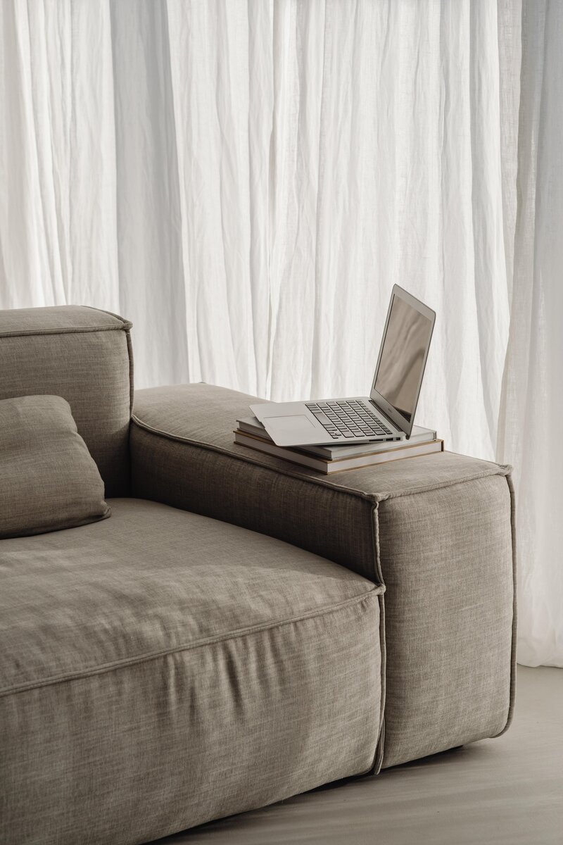 Macbook sitting on the arm rest of a sofa in front of a linen curtain.