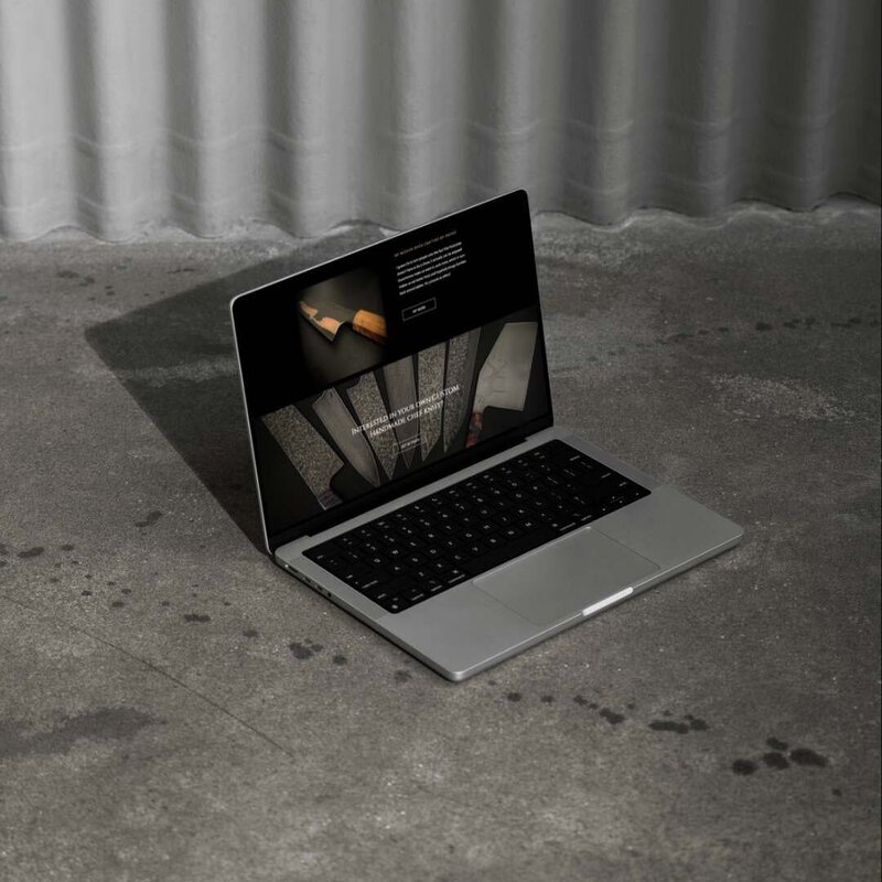 A laptop displaying a website for custom handmade chef knives, showcasing detailed images of various knife designs. The laptop is placed on a textured concrete floor with shadows cast, adding to the industrial aesthetic of the scene. The website features a dark theme with high-quality visuals of the knives.